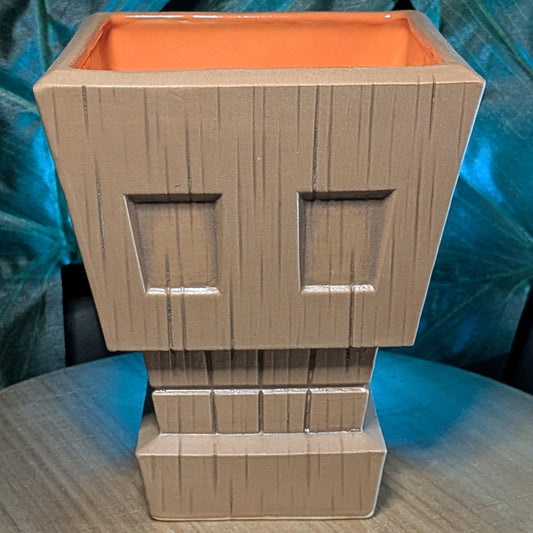 Wooden tiki mug with a stylized skull face, with an orange interior against a leafy background